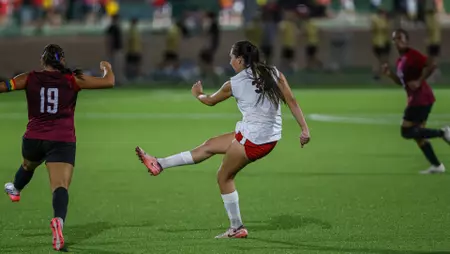 Female soccer player in a white jersey and red shorts kicks the ball as opponents in maroon chase on a bright green field.