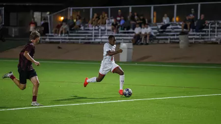 Soccer player in a white kit dribbles past a defender in maroon as they chase the ball on a well-lit grass field, with spectators seated in the background under stadium lights.