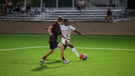 Soccer players contesting the ball on bright green turf at night; one in white with red trim shields the ball from an opponent in maroon and black shorts, bleachers in the background.