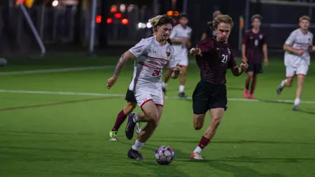 Two soccer players race for the ball on a lit artificial turf field at night, one in white and the other in maroon, with teammates in the background