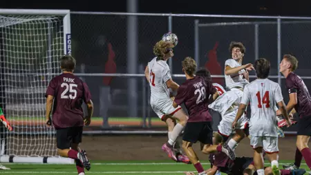 Soccer players in white and maroon jerseys jump around the goal as the ball descends toward the net during a night match, crowd blurred in the background.