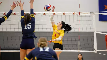 Female volleyball player in a yellow jersey (#4) spikes the ball over the net as two defenders in dark blue jump to block.