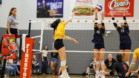 Female volleyball player in yellow jumps to spike the ball as two navy-clad blockers jump to block at the net; referee watches from the left in a gym.