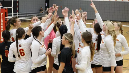 Girls' volleyball team in white uniforms huddles on the court, hands raised together in celebration or unity.
