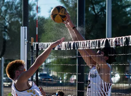Two outdoor volleyball players jump at the net, one spiking while the other attempts a block as the ball travels above the net edge in daylight.