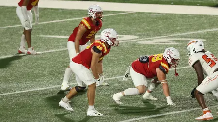 Football players in red and gold uniforms line up at the line of scrimmage on a green turf field.