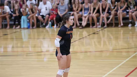 Female volleyball player in a black uniform with white arm sleeve and knee pads stands on an indoor court as spectators watch from the bleachers in the background.