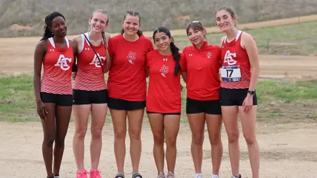 Six young women runners in red uniforms stand shoulder to shoulder on a dirt track outdoors, smiling; one wears bib 17.
