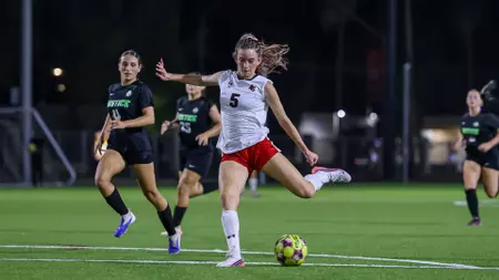Female soccer player in a white jersey (#5) kicks the ball on a night match, chased by opponents in dark uniforms on a green turf field.