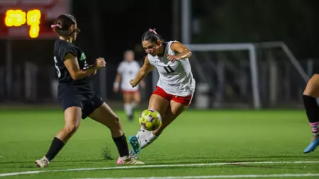 Female soccer player in white jersey and red shorts challenges the ball against a defender in black on a night game on green turf.