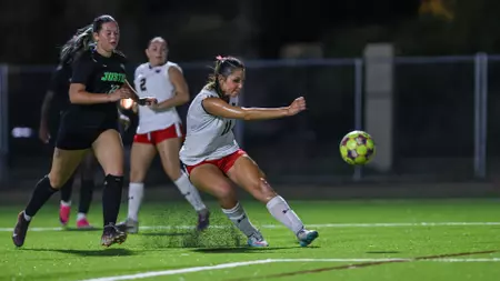 Female soccer players chase a bright green ball on a turf field at night; a player in white kicks the ball while others pursue.