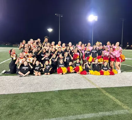Group of cheerleaders posing on a football field at night, holding red, yellow, and black pom-poms and smiling for a team photo.
