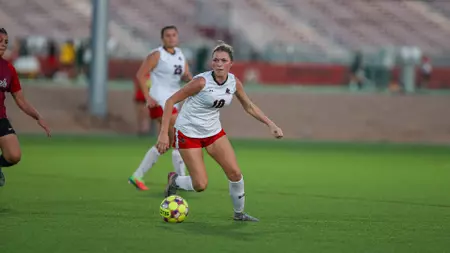 Female soccer player in a white jersey with red shorts dribbles a ball on a green field during a match.
