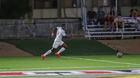 Soccer player in a white kit runs toward the ball on a lit turf field, with spectators watching from bleachers in the background at night.