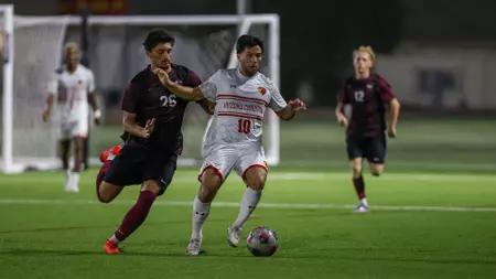 Two opposing soccer players chase the ball on a brightly lit field at night; maroon jersey 25 and white kit 10 contend for possession, with a goalie and another player in the background.