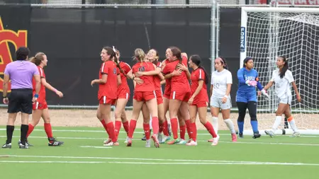 Women’s soccer teammates in red celebrate with a group hug on a green turf field near the goal after a match.