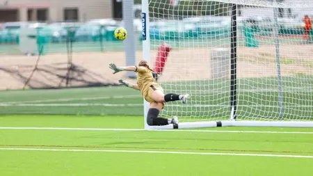 Soccer goalkeeper in a yellow jersey dives to block a high shot near the goal on bright artificial turf field.