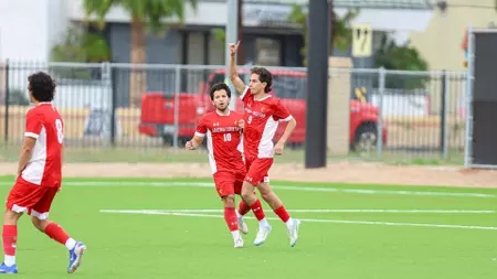 Three young soccer players in red uniforms celebrate on a green artificial turf field near a fence in the background.