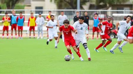 Soccer players in red and white chase the ball on a bright green field, with substitutes watching from the sideline.