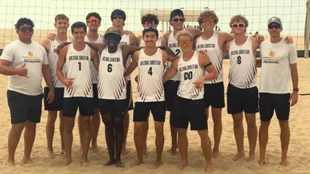 Group of young male beach volleyball players in white numbered jerseys posing together on a sandy court with a net behind them.
