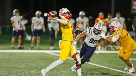 Quarterback in yellow throws a pass as a white jersey defender closes in on the tackle on a football field.