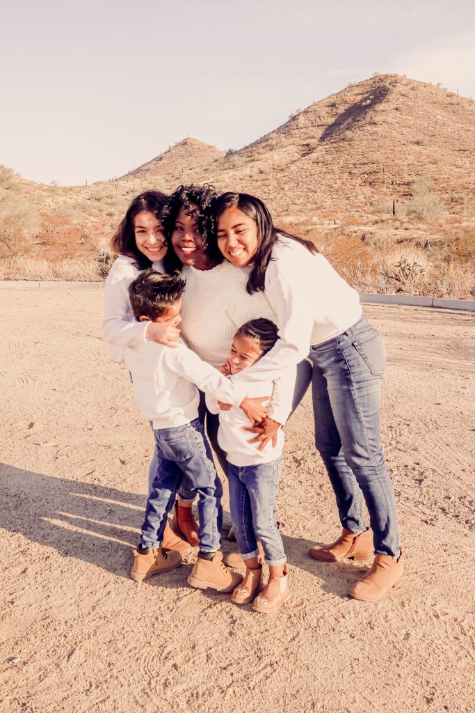 A family of five posing together in a desert landscape with rocky beige hills in the background, smiling and hugging.