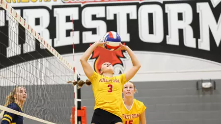 Volleyball player in yellow jersey (#3) jumps to set the ball at the net in a gym, with a teammate behind and a 'FIRESTORM' banner.