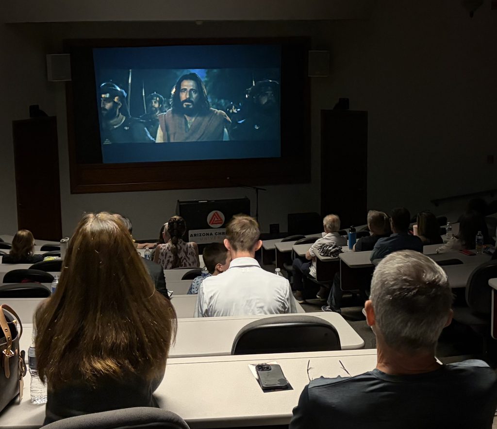 Audience seated in a dark theater watching a movie projected on a large screen displaying a bearded man in robes.