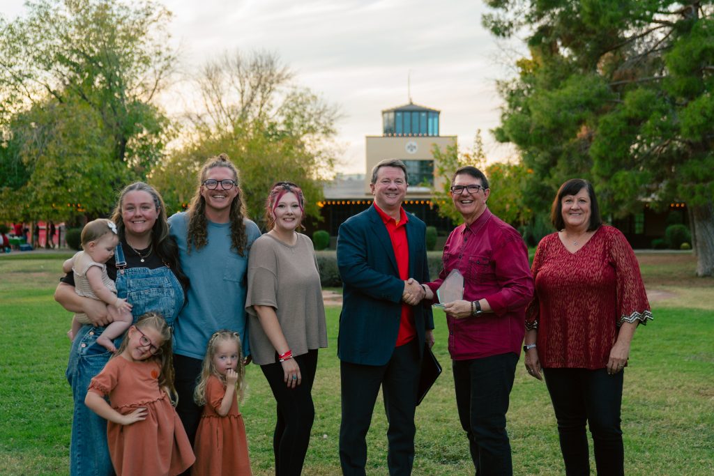 Group of adults and children posing outdoors on a lawn, smiling, with a building and trees in the background.